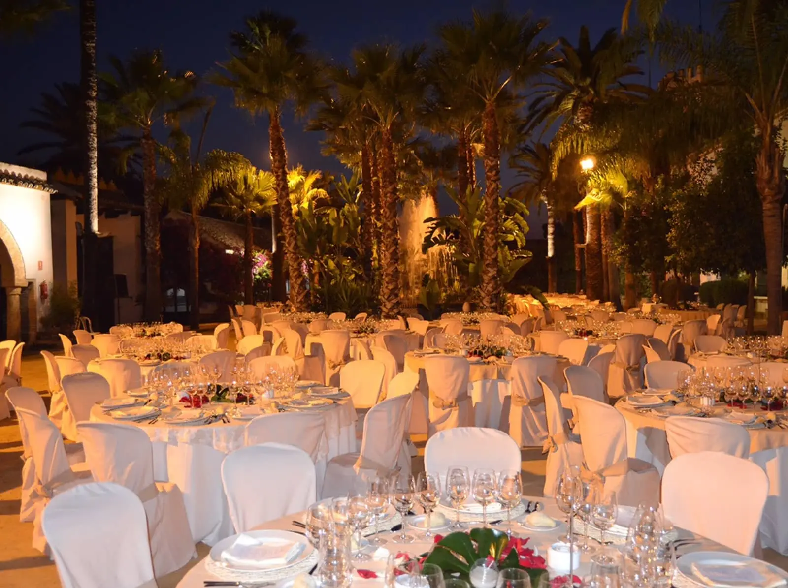 Mesas de banquete vestidas de blanco en el Patio Principal, de noche, bajo las palmeras iluminadas
